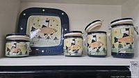 Front view of four folk art canisters and matching plate depicting fish, flag and fisherman. Plate has paint loss on bottom edge.