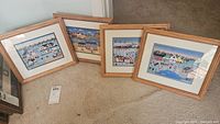 Four framed folk art prints by Colleen Sgroi laid on carpet, showing all frames and mats with colorful naive village scenes including houses, trees, and animals