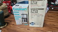 Photo of boxed Cuisinart 14 cup programmable coffee maker and two boxed West Bend 12-30 cup coffee urns on wooden floor.