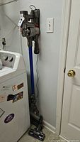Side view of Dyson stick vacuum mounted on wall next to laundry room washer, showing long blue extension wand and floor head.