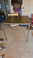 Side angle view of square folding card table with metal legs and brown wood-grain laminate top in worn condition.