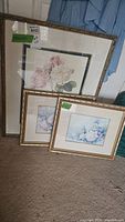 Three framed floral prints resting on carpet, two smaller prints with gold gilded frames stacked in front of a larger gold framed print with fabric matting featuring botanical flowers in pastel shades.