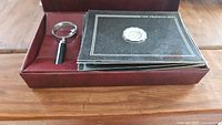 Burgundy box lined with velvet containing the miniature plates and a magnifying glass resting inside the box.