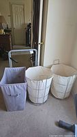Photo showing one rectangular gray canvas hamper, two round white canvas hampers with metal frames and handles, and a folding luggage rack with metal frame and black handles.
