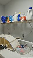 Photo showing shelf above the washer with detergent containers and hanging canvas storage partially visible