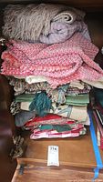 Stack of assorted linens including woven textiles with fringed edges in various colors such as red, beige, and green with tassels.