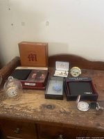 Wooden surface with Ford pocket watch under glass dome, Westclock pocket watch in plastic dome, Cardow desk clock, Tommy Hilfiger wallet box, Pronto Uomo boxed lapel pin, and two sets of cuff links, including a set with damage.
