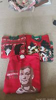 Three Christmas sweaters folded on carpet including cat, Santa Claus and Abraham Lincoln face designs.