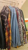 Photo showing multiple men's long sleeve button-down flannel and denim shirts hanging on hangers, various plaids and solid denim colors visible.
