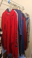 Seven men's sweaters, vests, and hoodies hanging on wire hangers on a closet rack. Includes red Nautica pullover hoodie, gray zip-up hoodie, and various knit sweaters and vests in red, blue, gray, and camouflage pattern.