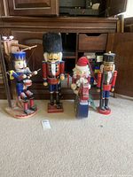 Four assorted wooden nutcrackers arranged on carpet in front of wooden cabinet; a Santa figure, a tall soldier with furry hat, a drummer with blue uniform, and a soldier with black helmet. Note damage to one nutcracker's foot.