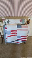 Two folded quilts on a wooden rack against wallpaper: one with traditional patchwork and one with flag motifs.
