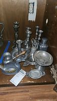 Photo showing pewter candlesticks of various sizes, pewter duck figurine with painted details, pipkin warming pot with wooden handle, pedestal dish, bell, and other silverplate items arranged on a shelf.