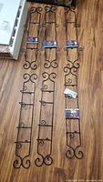 Three slender black wrought iron plate racks with scroll design, laid flat on wooden floor with tags attached to four of the racks.