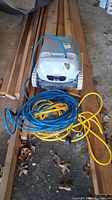 Overall front view of the white Dolphin Active 20 pool vacuum with yellow and blue hoses/coiled cords placed on wooden planks in a garage.