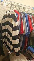 Photo showing multiple men's shirts hanging in a closet, including black and white striped rugby shirt and various colored t-shirts in red, blue, and gray shades.