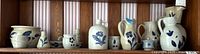 Full view of 12 beige Williamsburg pottery pieces decorated with blue hand-painted leaf and floral motifs arranged on wooden shelf with striped wallpaper background.