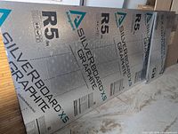 Full and partial sheets of Silverboard XS Graphite foam insulation board leaning against a wall, showing front reflective foil-faced surface with printed text and specs.