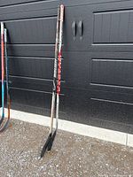 Two adult hockey sticks standing side-by-side against a black garage door, one mostly black and red (True A6.0 brand) and the other blue and black (Sher-Wood brand), both with taped blades for right-handed shooters.