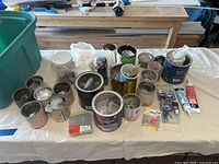Photo showing multiple metal cans, jars, and retail packages on a table, each containing different types of screws, nails, fasteners, and adhesives.