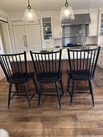 Set of three black metal counter stools placed in front of a kitchen island with white cabinetry and wooden floor, viewed from the back showing vertical slat metal backs.