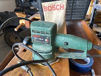 Bosch PEX 115 A random orbit sander on wooden surface with Bosch dust collection bag behind it.