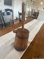 Front angle view of the wooden butter churn on a dining table with chairs in background.