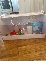White chick brooder box with clear plastic lid and two heat lamps inside, includes feeders, water dispensers, and puppy pads inside.