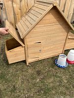 Side view of wooden chicken coop showing hinged nesting box open and feeder and waterer beside it on grass.
