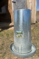Galvanized metal cylindrical chicken feeder with tray at bottom, outdoors on grass near a wooden coop.