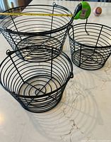 Three black wire egg baskets shown with measuring tape for scale on a light-colored surface.