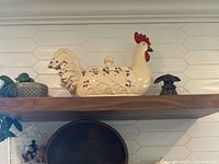 Side view of cream ceramic chicken crock with red comb and black speckled accents on a shelf.