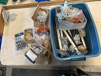 Overall view of assorted hardware items in a large blue plastic bin, several smaller cardboard boxes and loose items on a table.