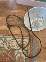 Heat lamp lying on carpeted wooden floor, showing white reflector with protective grid and black power cord with switch.