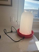 Photo of a heated poultry waterer placed on a kitchen counter near window with daylight. Shows translucent white water reservoir mounted on a red base with black power cord coiled nearby.