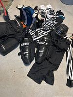 Full collection of adult large hockey refereeing equipment including protective shin guards, pants, gloves, elbow pads, striped jersey, helmet cage, protective cup, and hockey bag shown laid out on floor.