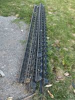 Nine black plastic landscaping paver edging pieces laid on grass and gravel, showing full length and pointed stakes for ground insertion.