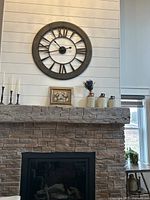 Photo of large round wall clock on a white paneled wall above a stone fireplace mantel with decorative items around it.