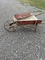 Side view of an old wooden wheelbarrow with red faded paint and metal spoked wheel on gravel background.
