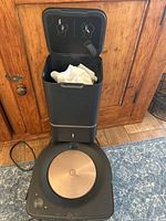 Front view of Roomba robot vacuum docked in Clean Base station with lid open showing disposal bag