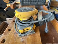 Yellow and black Dewalt 1/4 sheet sander on wooden surface with attached power cord coiled around it.