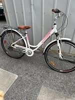 Full side angle of white and pink woman’s bike showing frame, saddle, rear rack, tires and chain.