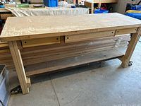 Full view of the wood workbench showing the solid wood top, 3 drawers and lower shelf, positioned on a concrete floor with various workshop items around.