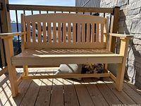 Yellow plastic wood garden bench with slatted backrest and solid armrests shown outdoors on wooden deck near stone wall.