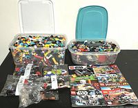 Two large plastic containers filled with assorted loose LEGO bricks in multiple colors, several sealed small bags with LEGO pieces, and multiple LEGO instruction manuals laid flat on table.