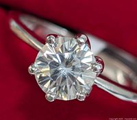 Front angle close-up revealing round brilliant moissanite held by six prongs on white gold band with red background