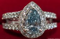 Close-up view of pear-shaped light blue lab diamond surrounded by small clear stones on silver band.