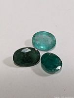 Three oval-cut genuine emerald gemstones placed on a white surface showing natural inclusions and varying green shades