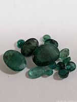 Genuine loose emerald gemstones in various sizes and cuts laid on white background. Oval and round shapes visible with deep green color and natural internal characteristics.