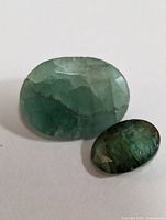 Image displaying two oval faceted emerald gemstones on a white background, showing their natural color and inclusions.